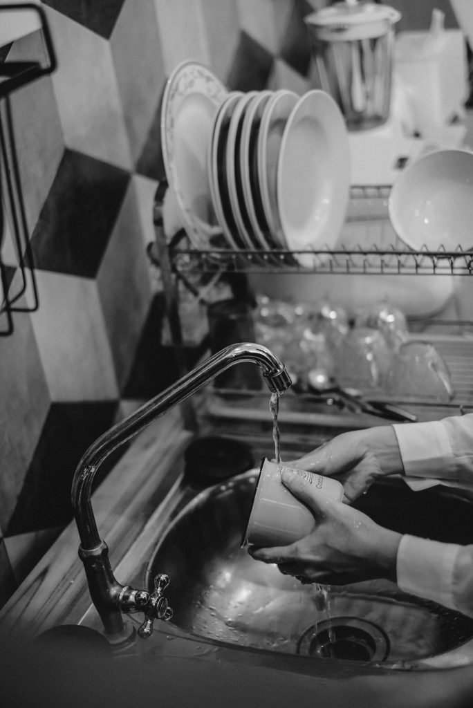 Black and white image of hands washing a mug at a kitchen sink with stacked crockery.
