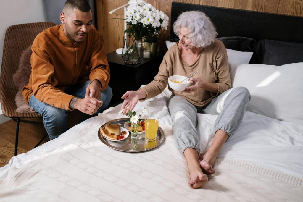 Elderly woman and young man enjoying breakfast in a cozy bedroom setting.
